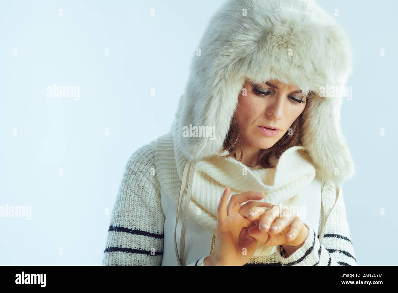 concerned modern female in white striped sweater, scarf and ear flaps hat having cold allergy against winter light blue background. Stock Photo