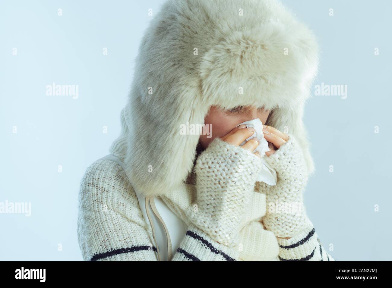sad modern 40 years old woman in white striped sweater, scarf and ear flaps hat wiping nose with napkin on winter light blue background. Stock Photo