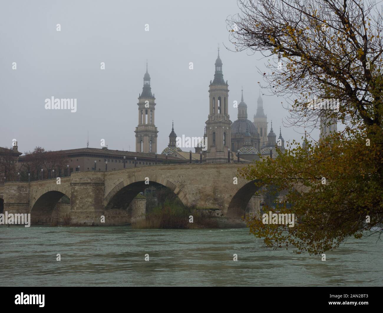 Stone bridge and the Basilica of Our Lady of The Pillar as seen from the Ebro river on a foggy day in winter time. Zaragoza, Spain. Stock Photo