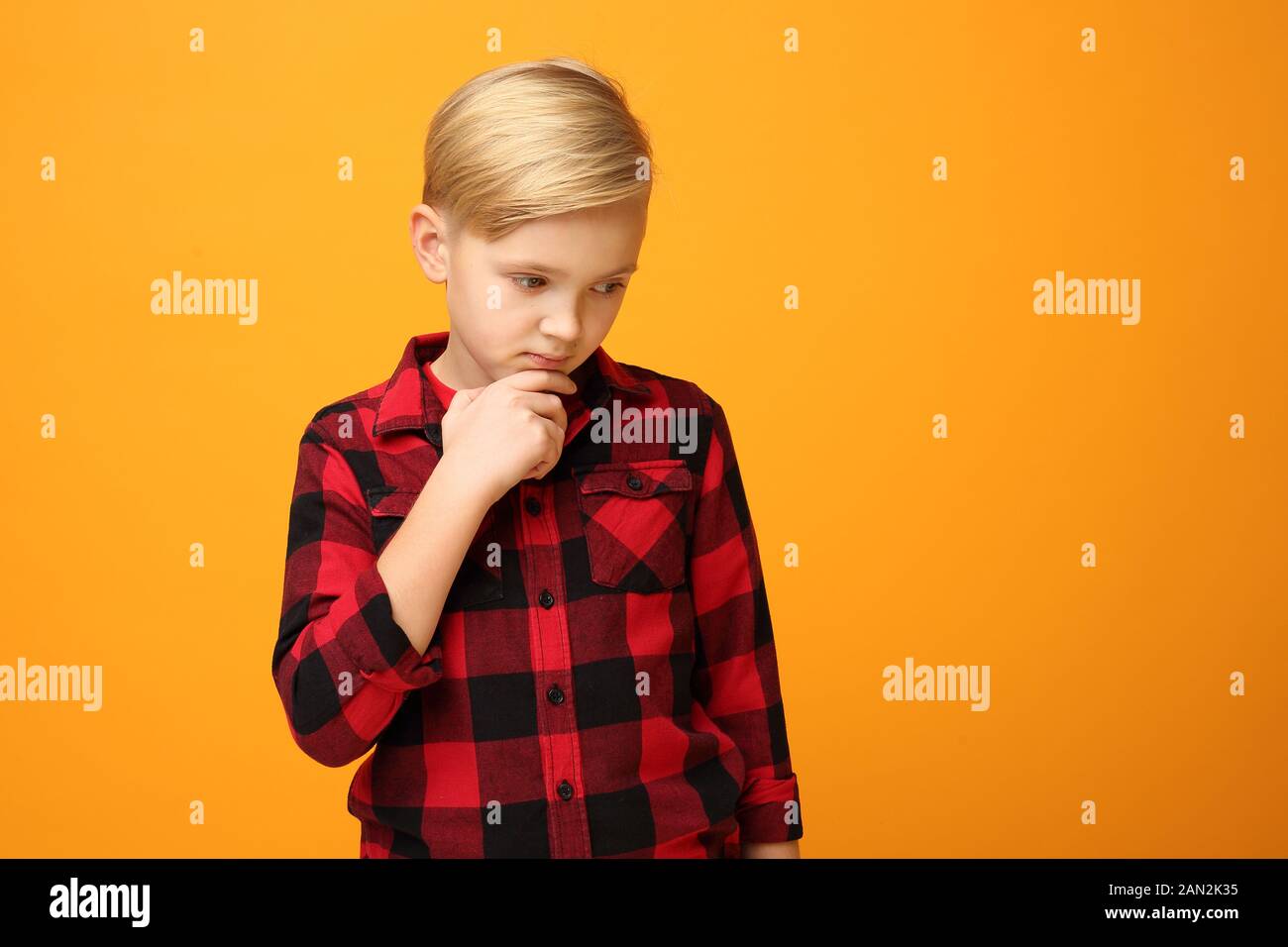Pensive focused young boy, child emotions. Young handsome smiling caucasian boy in the red shirt on the yellow background. Horizontal, straight on. Stock Photo