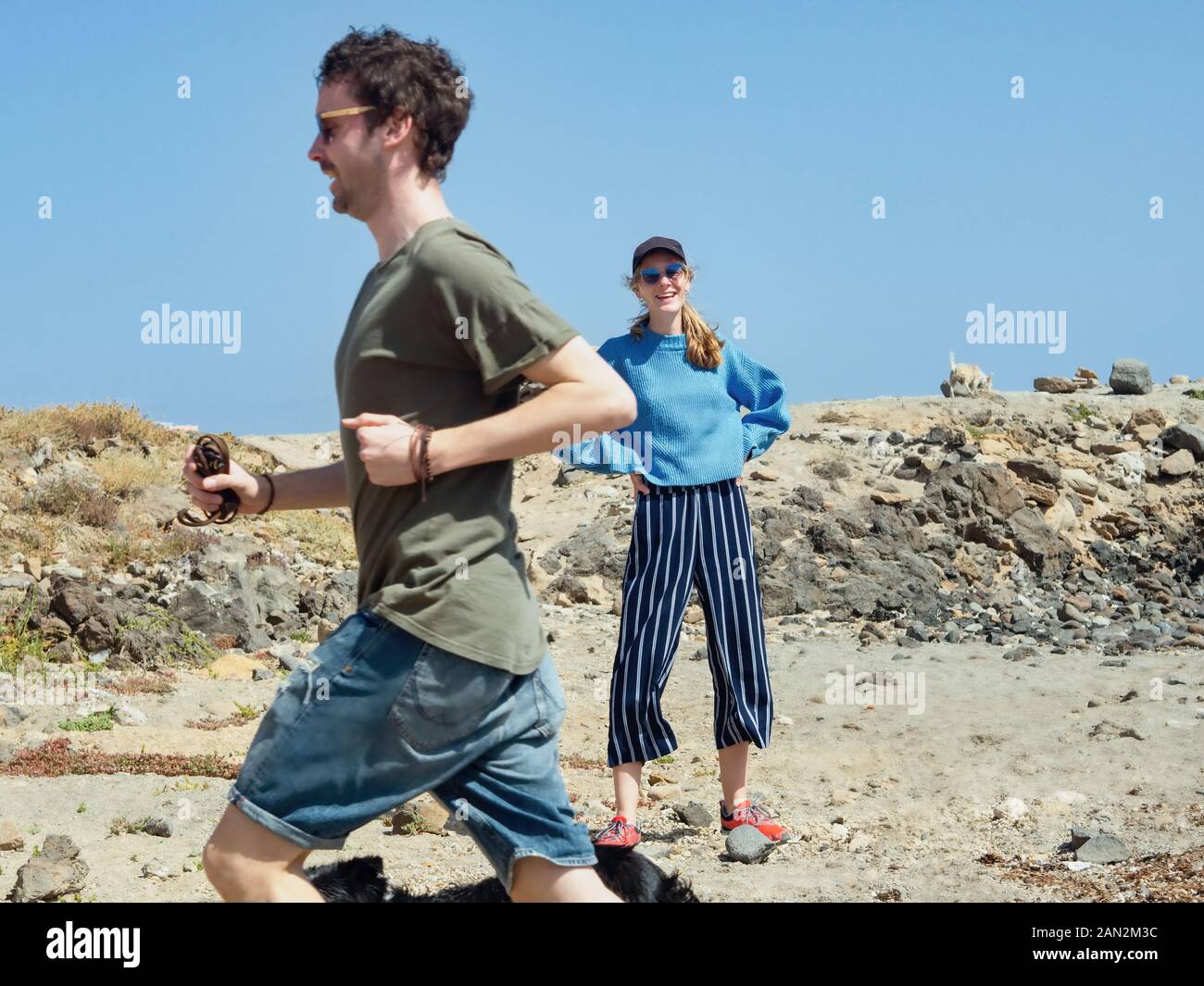 A laughing young woman with a black cap watches her boyfriend who has started a race with her dog. The young man runs past her. Very blue sky and sunn Stock Photo