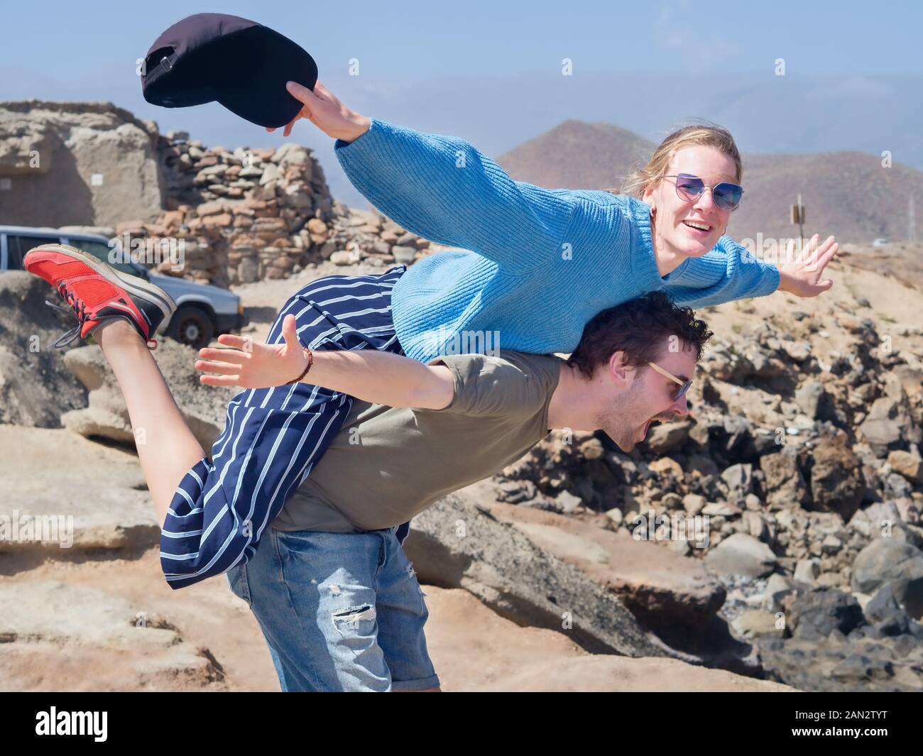 Two very young people, a pair of lovers, out in nature. He carries her on his back with arms outstretched in the position of an airplane. Both laugh a Stock Photo