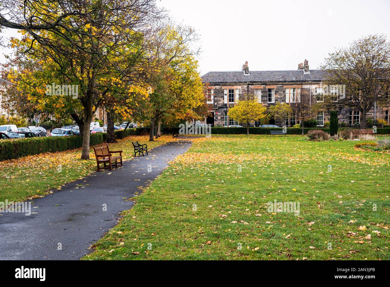 Public park with a paved path lined with wooden benches surrounded by tradtional stone terraced houses on a cloudy autumn day Stock Photo