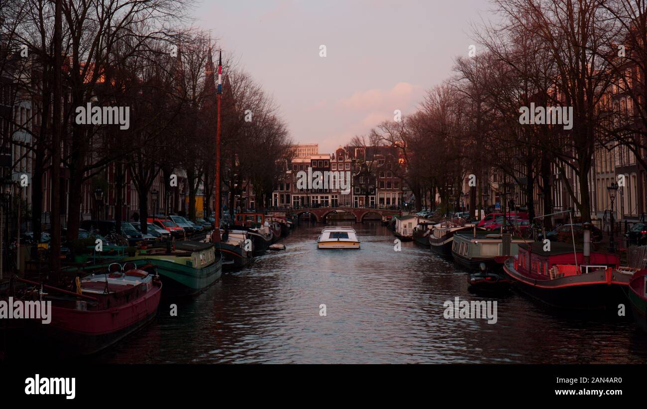 Boat cruises along a canal in Amsterdam with boats parked on both sides of it as it approaches a bridge Stock Photo