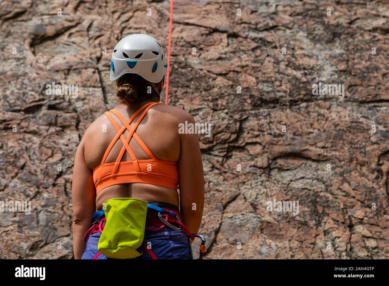 A close up and rear view of a healthy caucasian woman wearing orange vest, safety hat, harness and climbing gear during lead climbing a rock face Stock Photo
