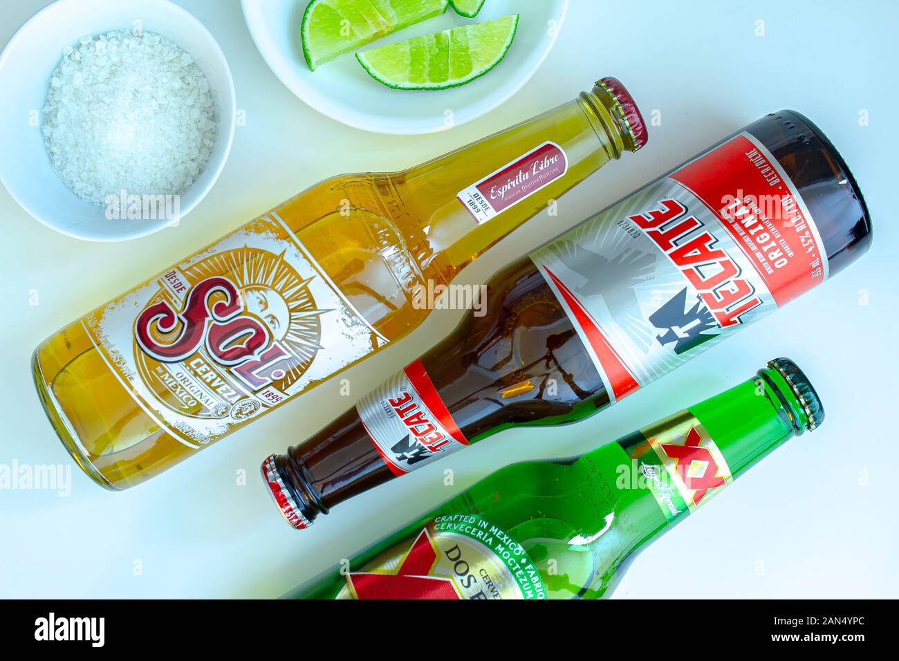 Bottles of Mexican pale and dark lager beers call: Sol, Tecate and Dos Equis on a white table with limes and salt on the side Stock Photo