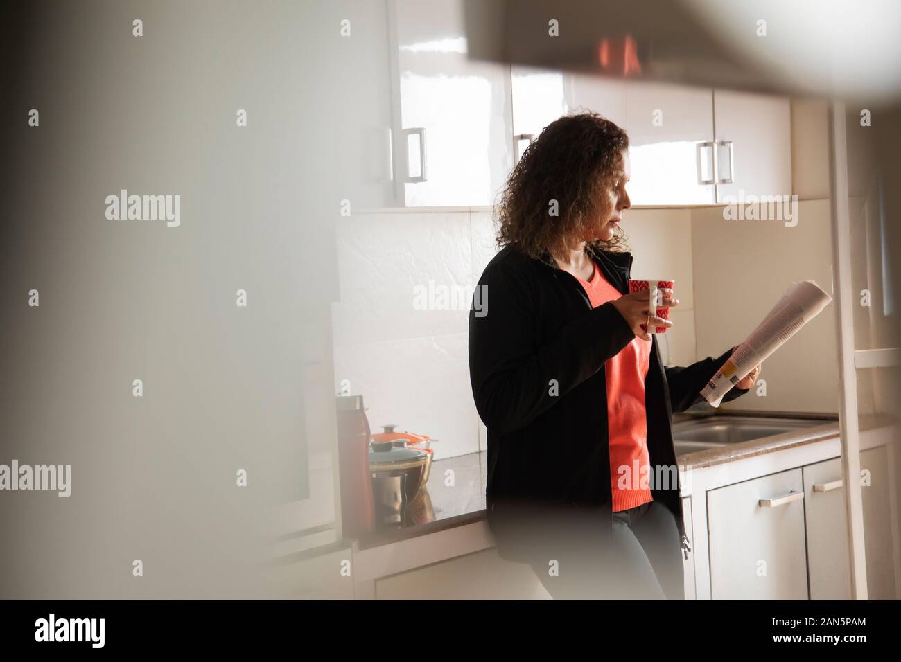 Woman drinking coffee while reading a newspaper in the kitchen at home. Stock Photo