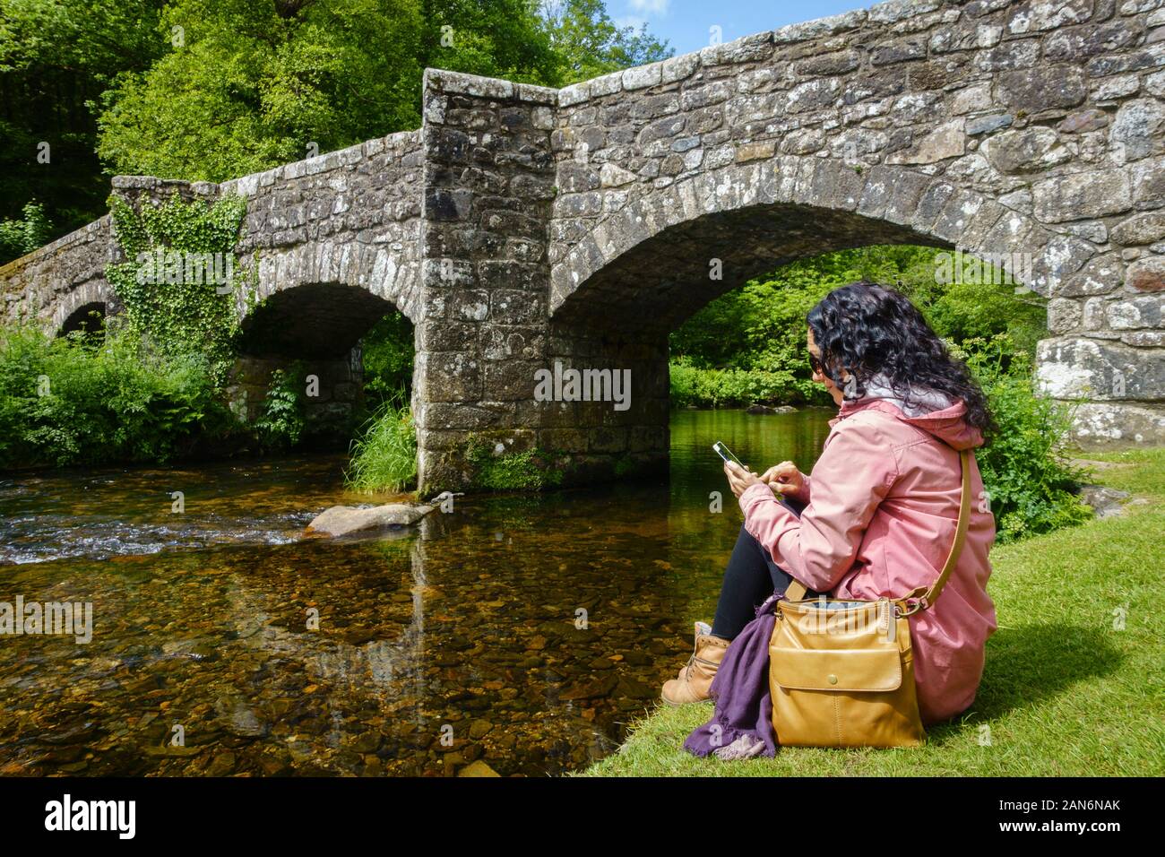 Brunette white woman using her mobile while sitting by a river and antique stone bridge. Always connected concept. Stock Photo