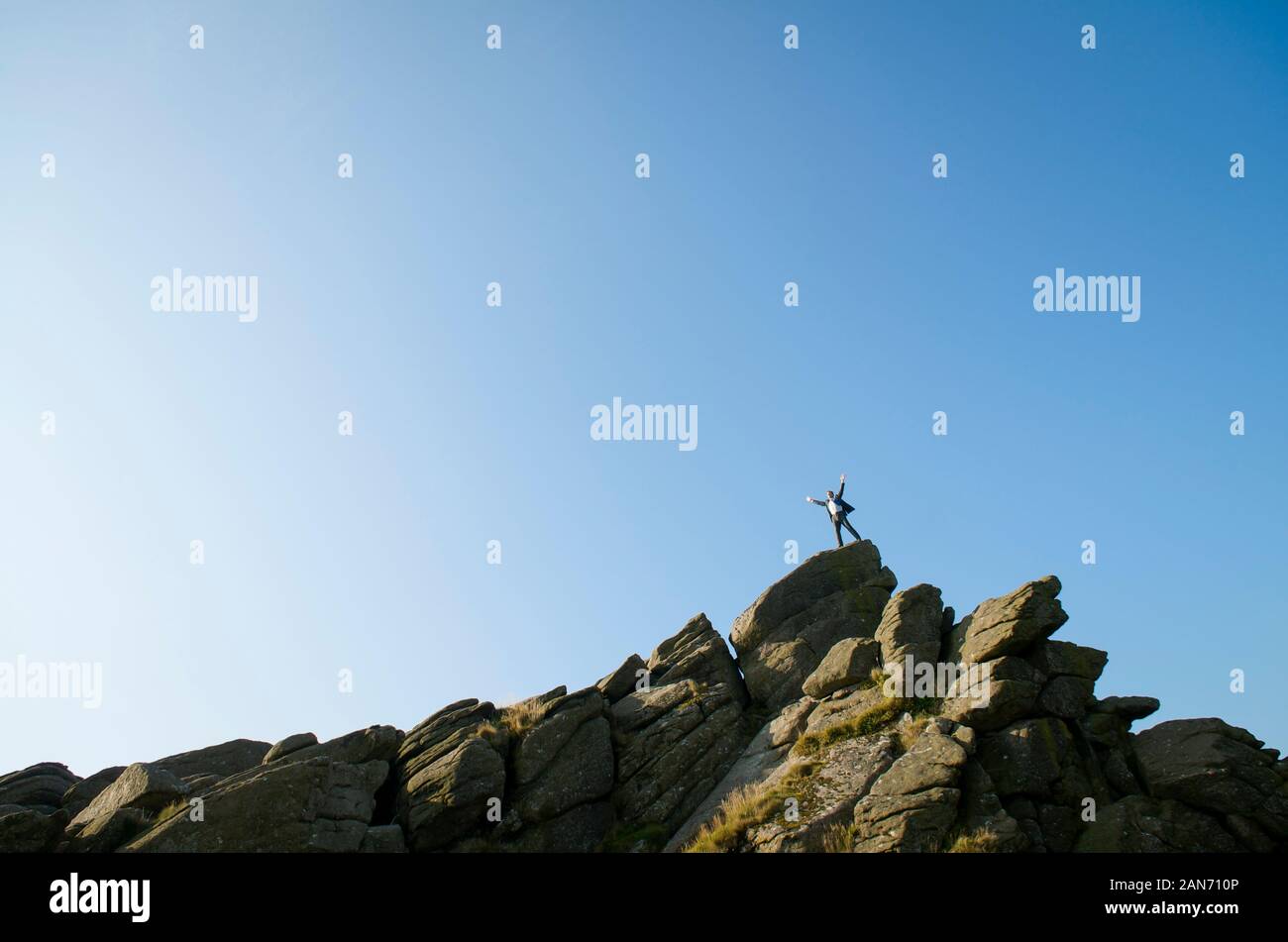 Distant businessman standing on the top of a rocky mountain peak celebrating in blue sky copy space Stock Photo