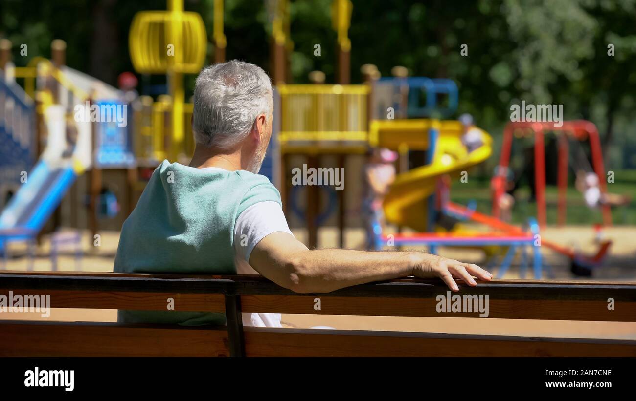 Male pensioner sitting on bench and watching grandkids on playground, family Stock Photo
