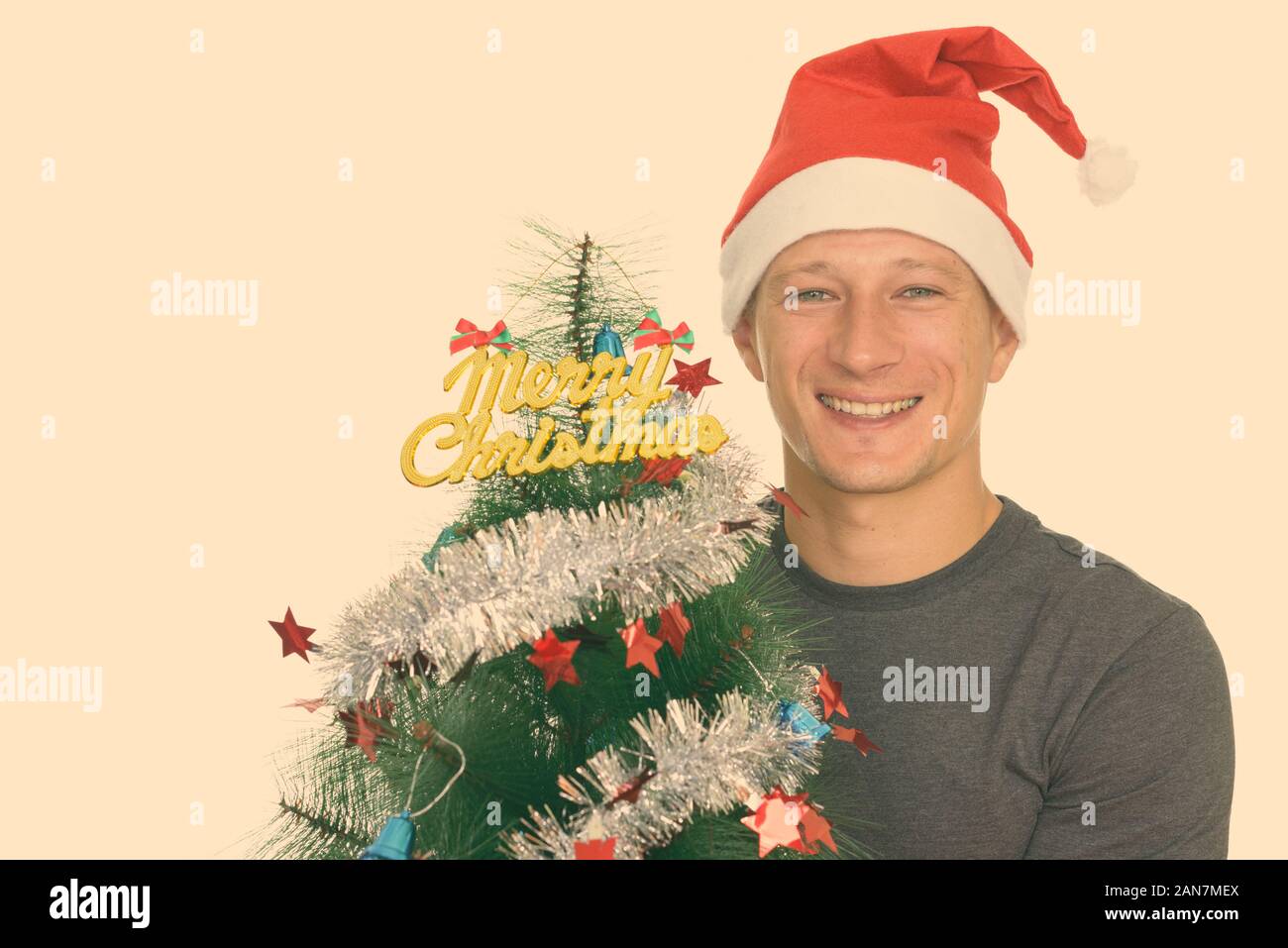 Studio shot of young handsome Caucasian man ready for Christmas holding Merry Christmas tree Stock Photo