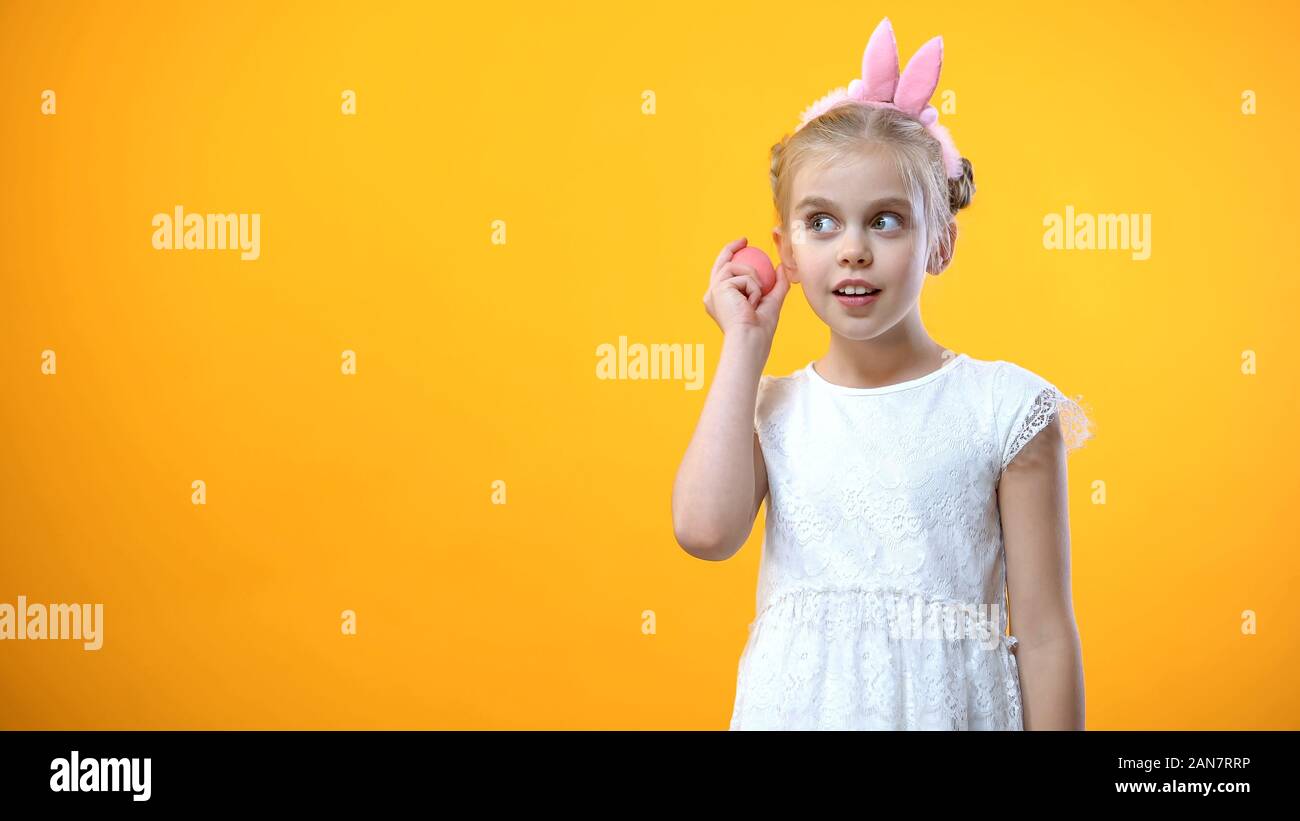Cute girl in white dress listening to pink egg on yellow background, festival Stock Photo