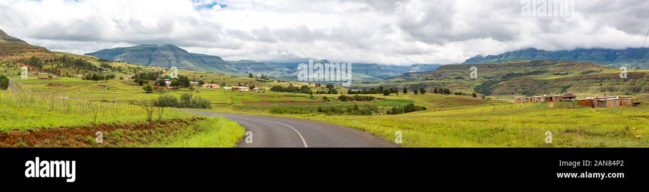 Panorama with a street leading to the mountains of Maloti Drakensberg, little village at the roadside, cloudy day, South Africa Stock Photo