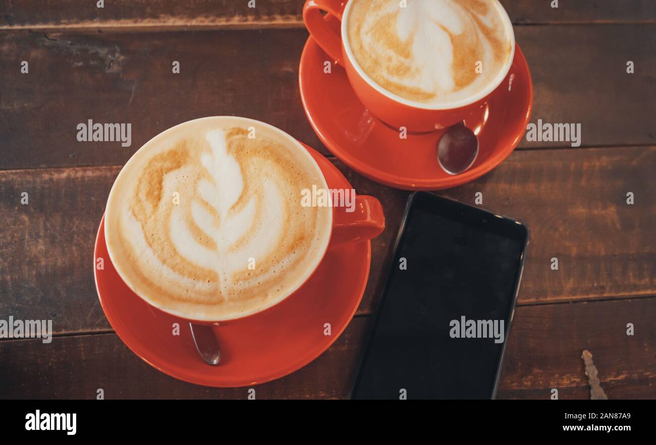 Two red cups of coffee with latte art on wooden desktop with the smartphone. Table in cafe. Top view. Stock Photo