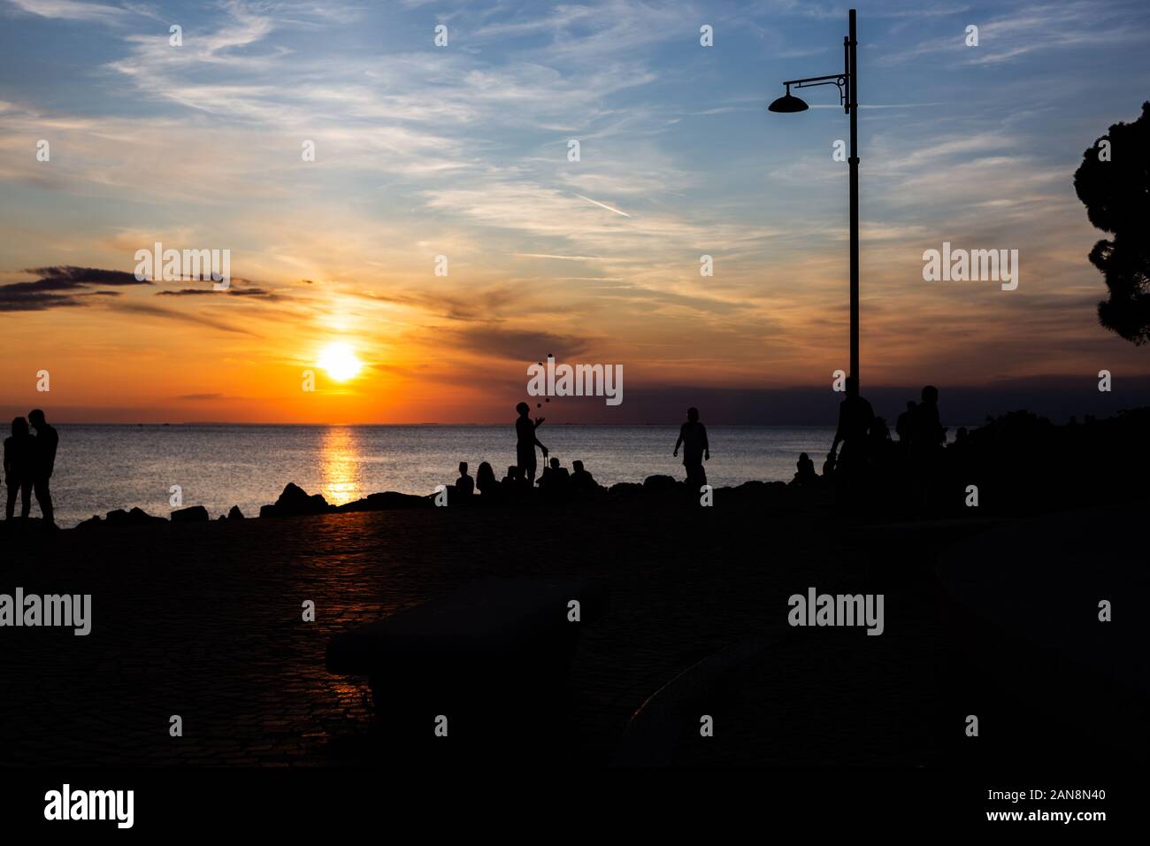 Dramatic and beautiful sunset at the coast line of the mediterranean sea in trieste Italy with silhouettes of a couple and people sitting on the beach Stock Photo