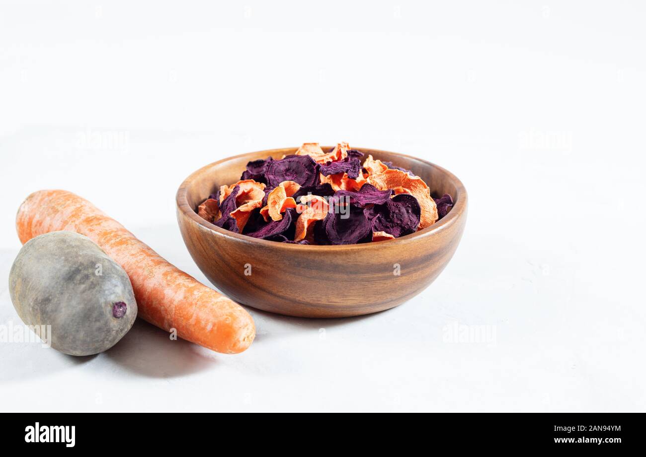 Dried carrot and beet chips in a wooden bowl and raw carrots, beets on a white concrete background. Organic natural food. Copy space. Stock Photo