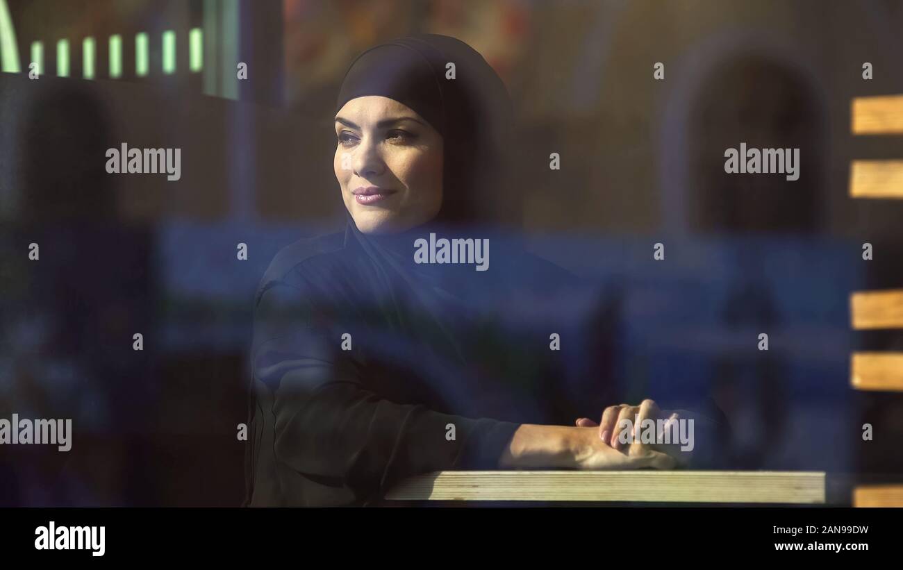 Smiling arab woman looking at city through window sitting in cafe, leisure Stock Photo