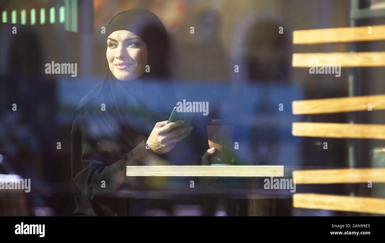Beautiful Muslim lady holding coffee and smartphone in hands, sitting in cafe Stock Photo