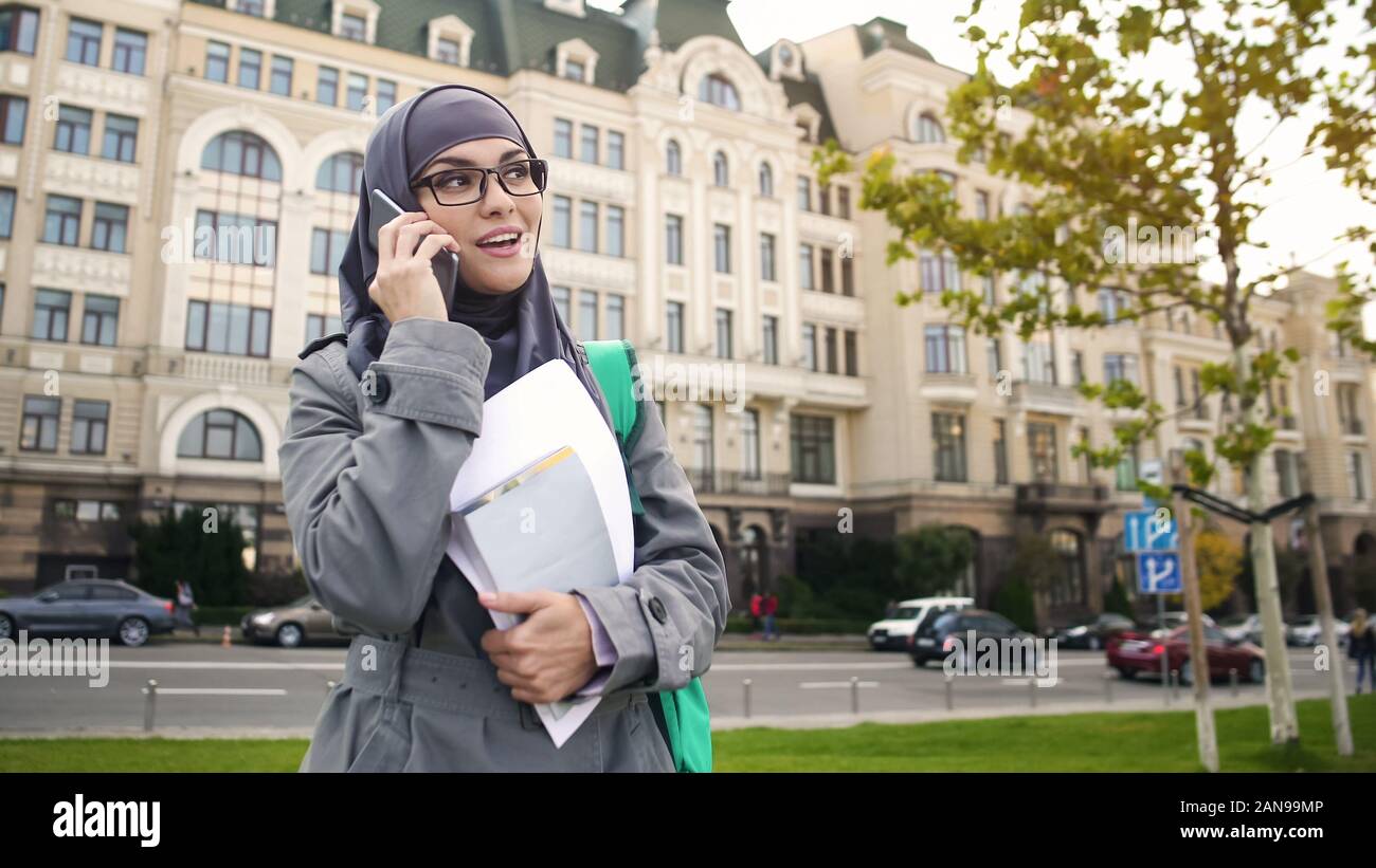 Self-confident smiling Muslim female student talking on phone standing on street Stock Photo