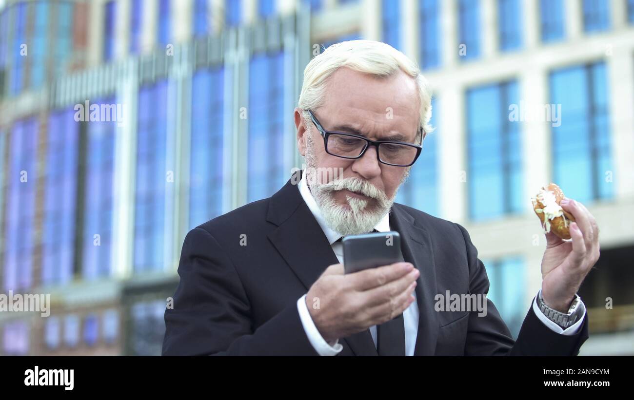 Elderly male in suit eating burger checking e-mail, stressful job, lack of time Stock Photo