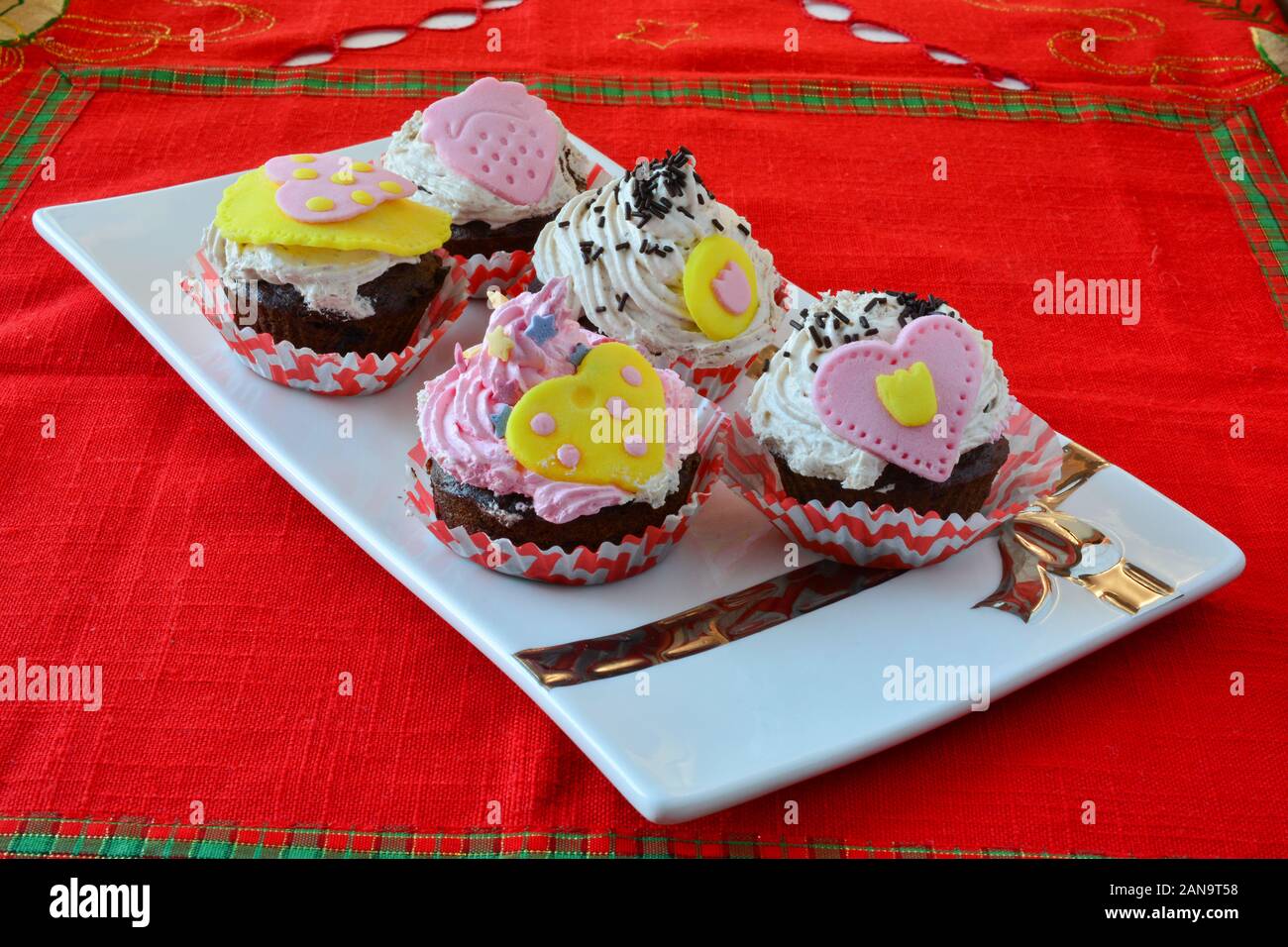 Love muffins, colorful velvet cupcakes decorated with marzipan hearts served in white ceramic saucer on table covered with red table cloth, side view Stock Photo