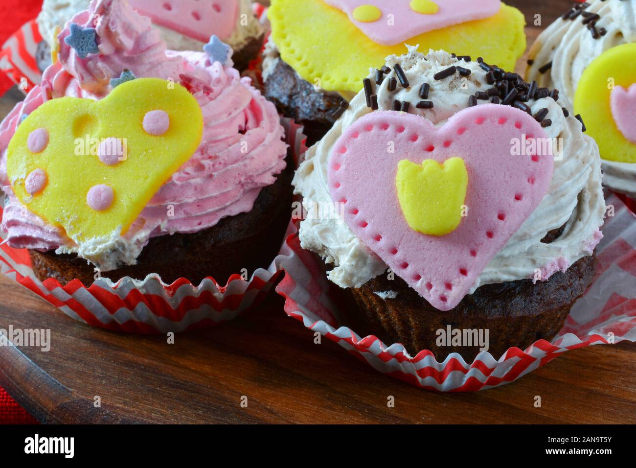 Valentine love muffins, colorful cupcakes decorated with marzipan hearts served on wooden round saucer over red table cloth, close up view Stock Photo