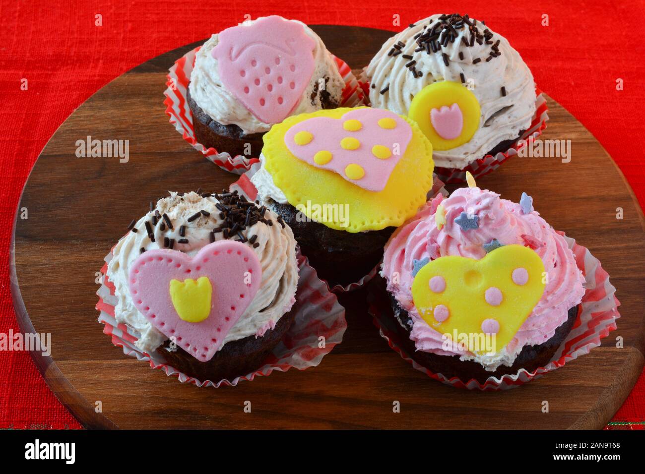 Love muffins, colorful cupcakes decorated with marzipan hearts, served on wooden round saucer over red table cloth, side view Stock Photo