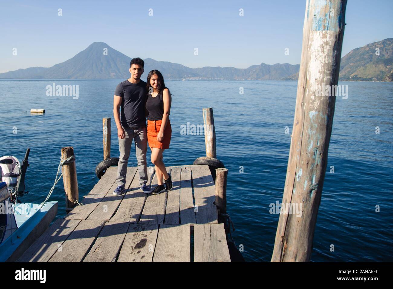 Traveling couple standing at the pier of Lake Atitlan smiling at the camera. Stock Photo