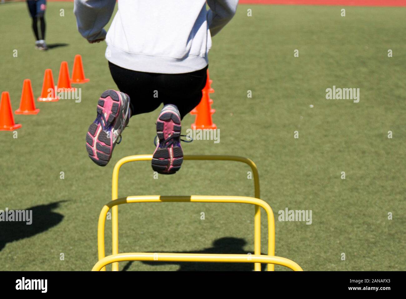 A track and field athlete jumps over a yellow hurdle while doing agility drills on a green turf Stock Photo