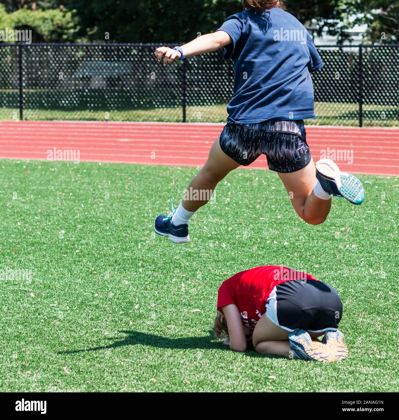 A high school track and field athlete hurdles her teammate during a game of leap frog at a fun practice day in the summer. Stock Photo