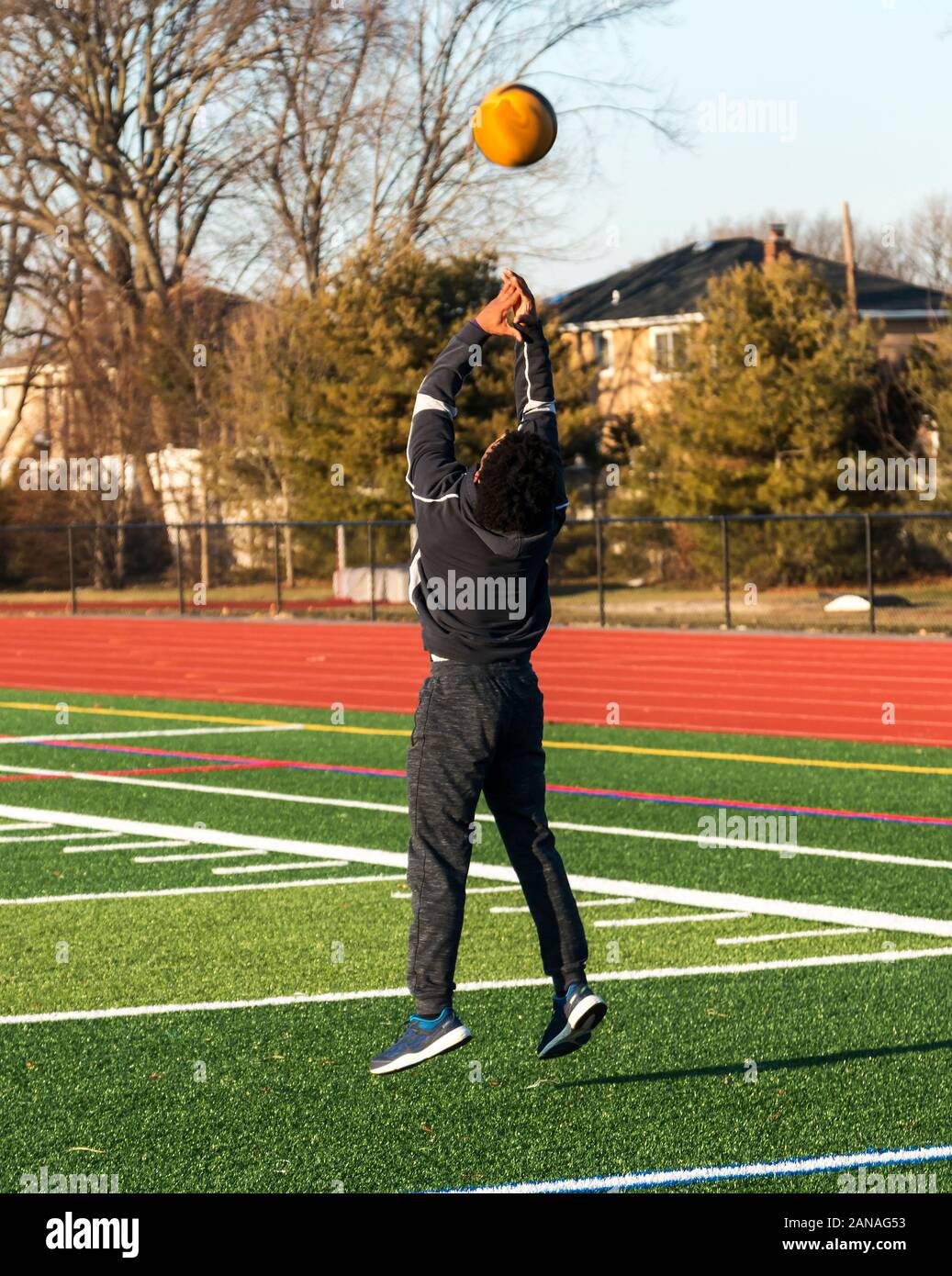 A high school track and field athlete is jumping in the air while throwing a medicine ball over his head backwards on a green turf field. Stock Photo
