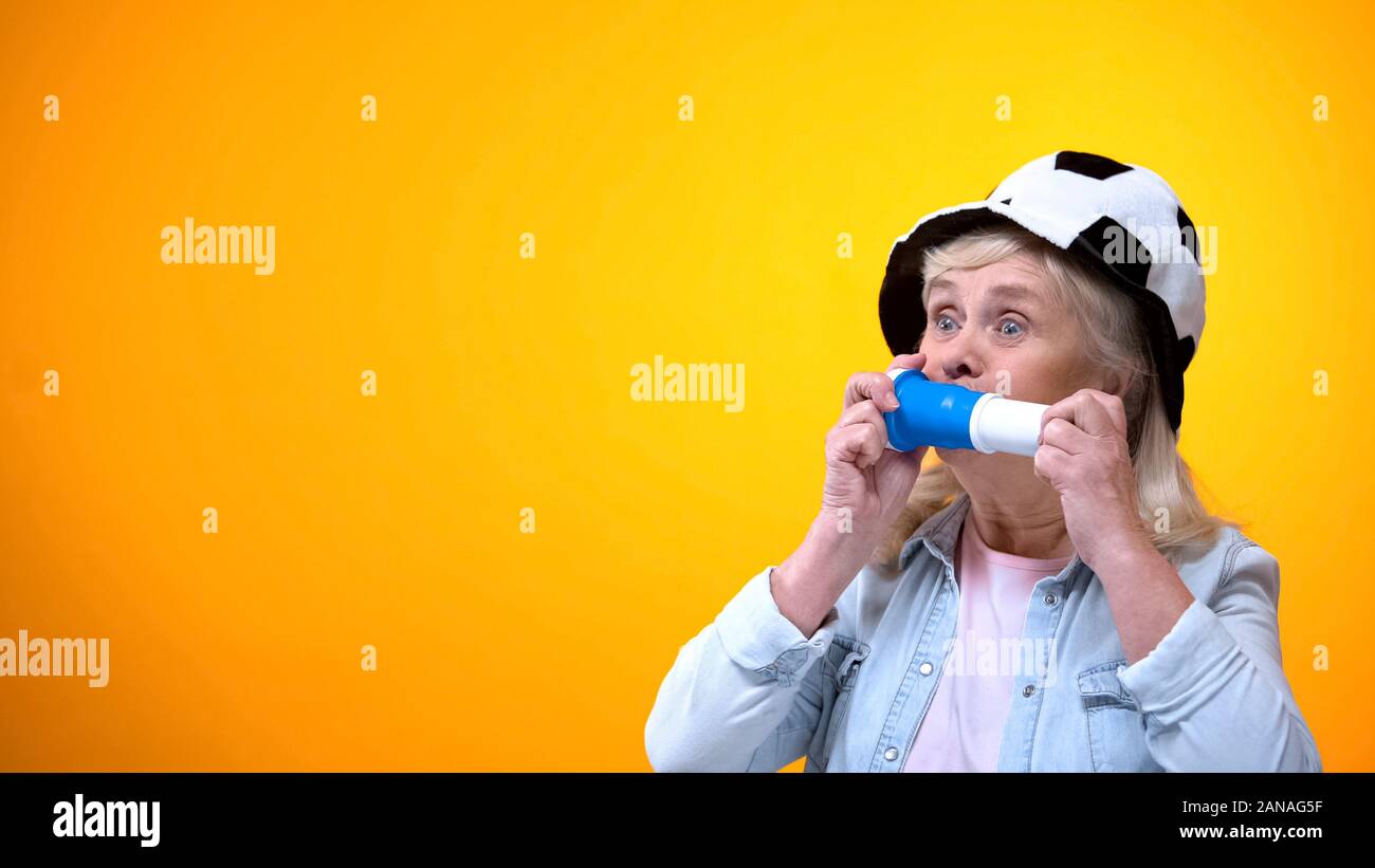 Joyful elderly lady in football fan hat blowing horn, sport betting, active life Stock Photo