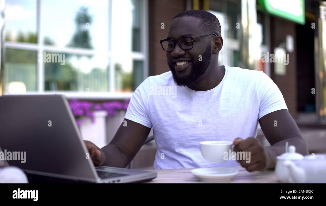 Happy afro-american man texting with relatives on laptop, sitting in restaurant Stock Photo