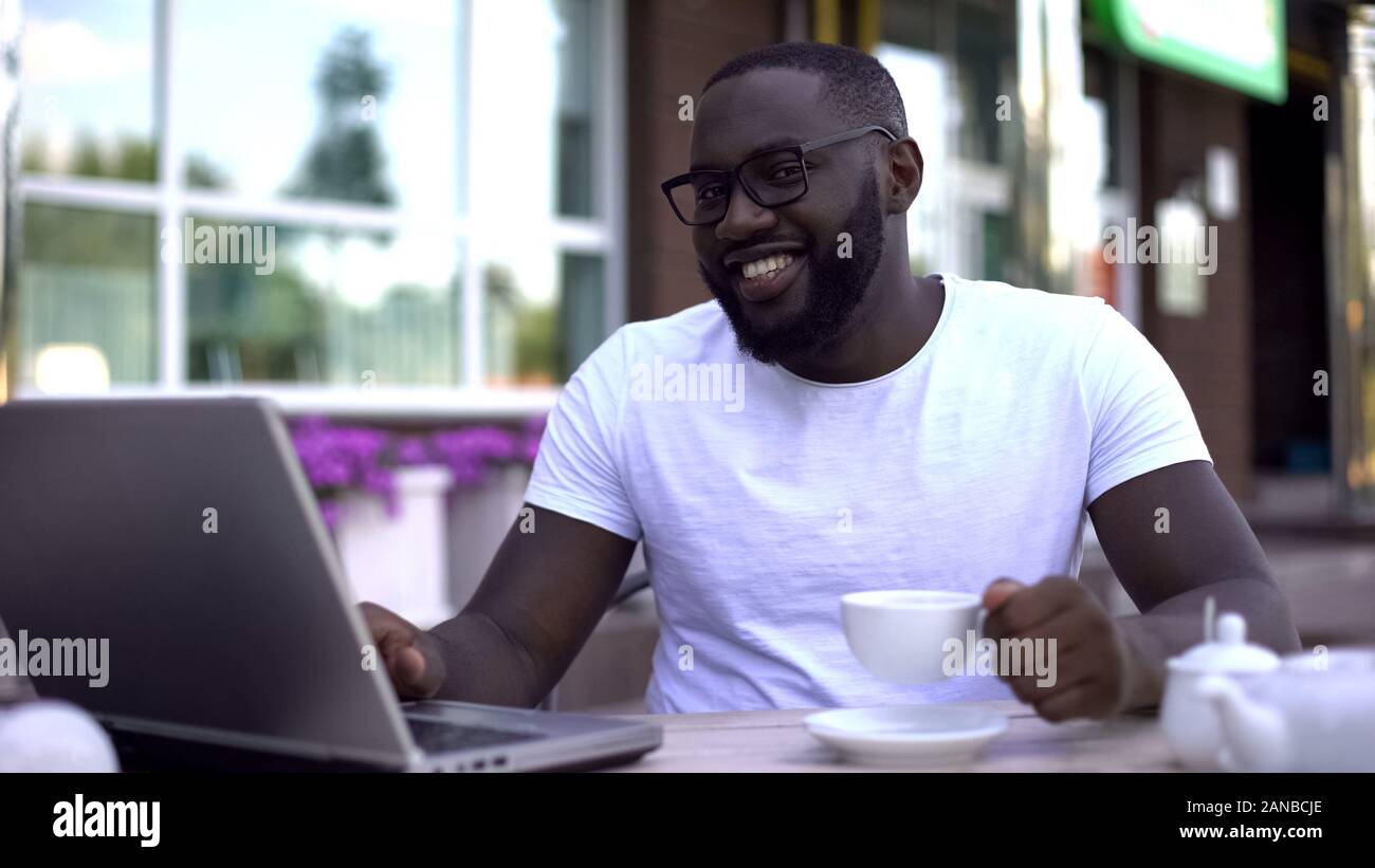 Smiling freelancer working on laptop drinking coffee and looking at camera Stock Photo