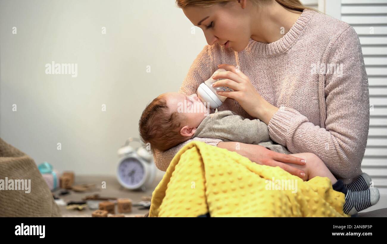 Young lady feeding baby boy, bottle with infant formula, putting kid to