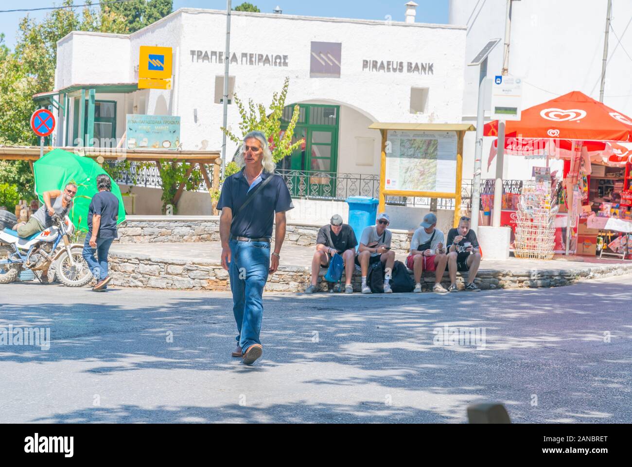 Filoti Naxos - August 13 2019; Middle aged Greek man with long grey hair  crosses street where four tourists take a break sitting on pavement behind  o Stock Photo - Alamy