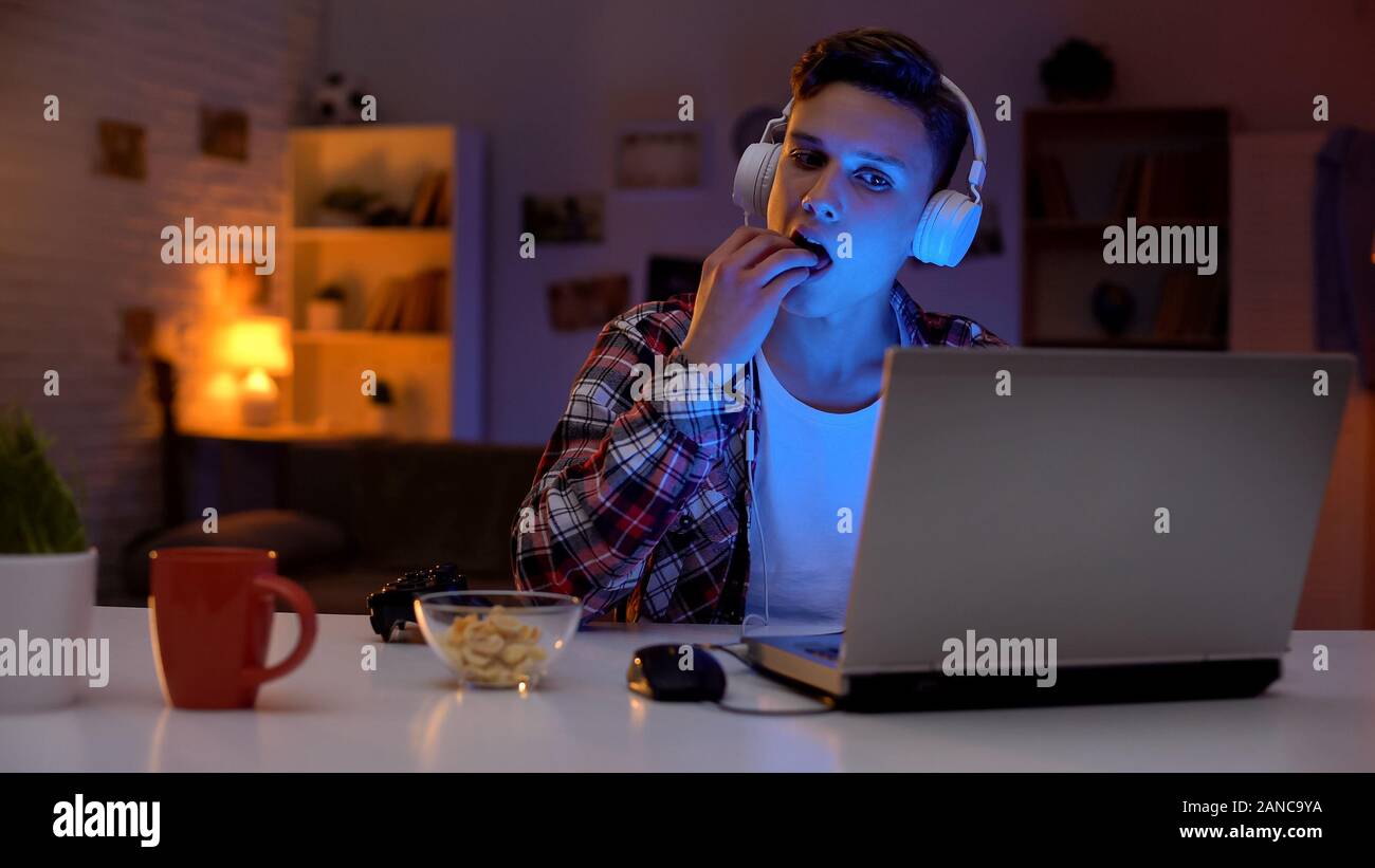 Excited teenage boy playing computer game and eating snack, leisure ...