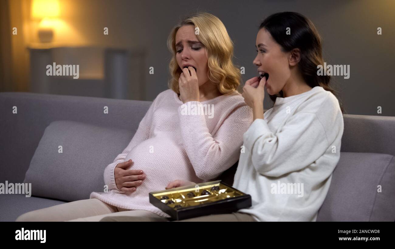 Woman sitting with upset pregnant friend, eating chocolate candies