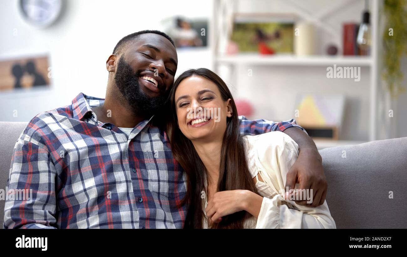 Nice multiethnic couple smiling and sitting on sofa at home, having fun together Stock Photo
