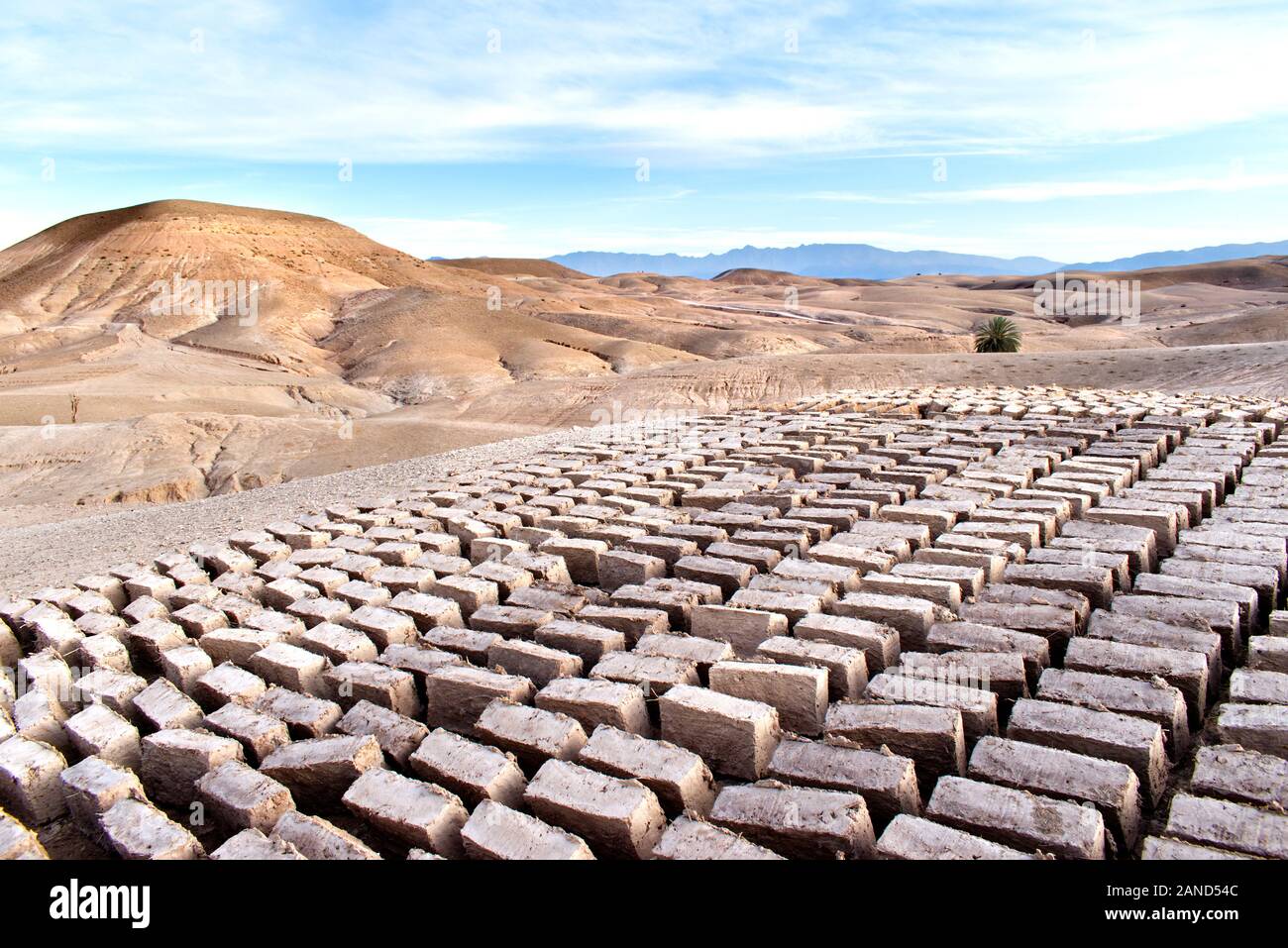 Mud bricks drying in the Moroccan desert with sand dunes and the foothills of the Atlas Mountains in the background. The landscape is under a blue sky Stock Photo