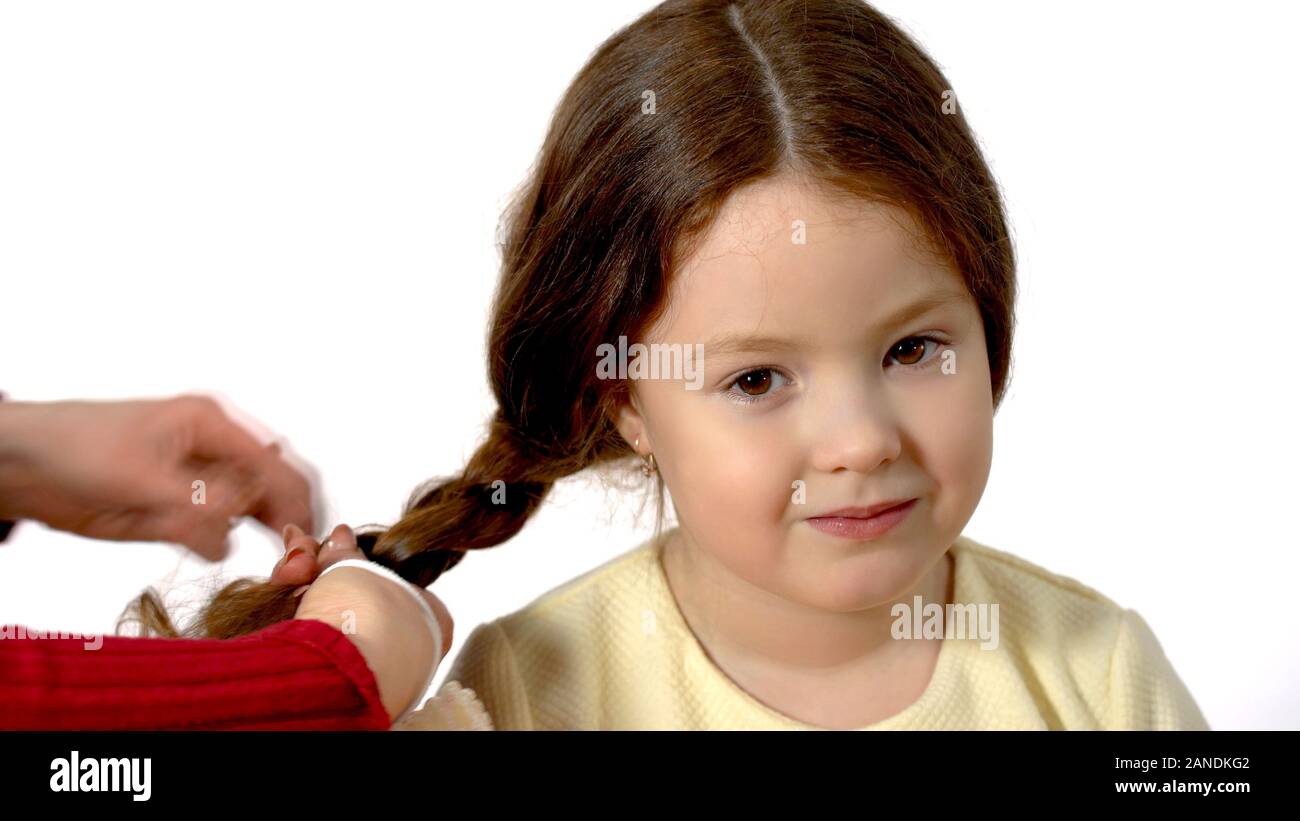 Mom braids the pigtail of her daughter. Studio photography on a white background. A 6 year old cute girl with brown hair looks at the camera and smile Stock Photo