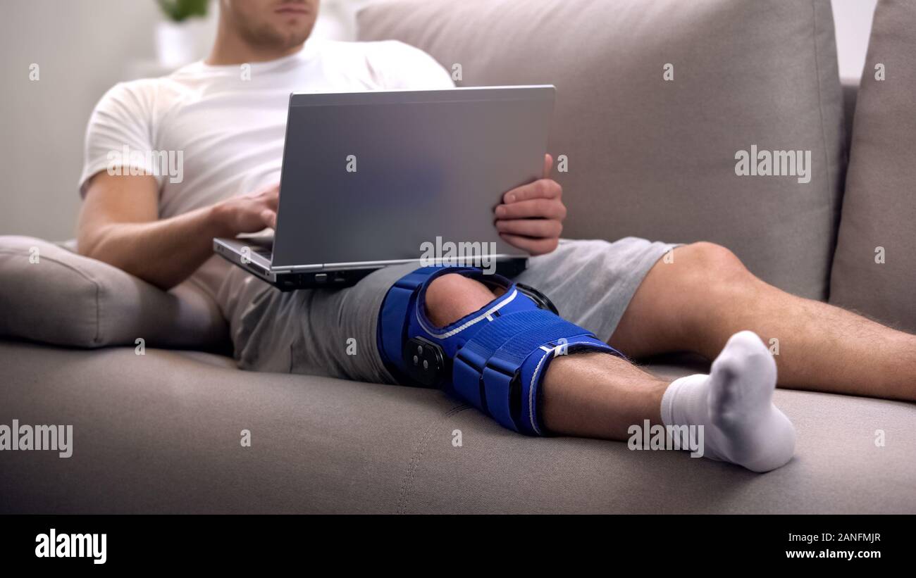 Man in arthritis knee brace working on laptop at home rehab period and
