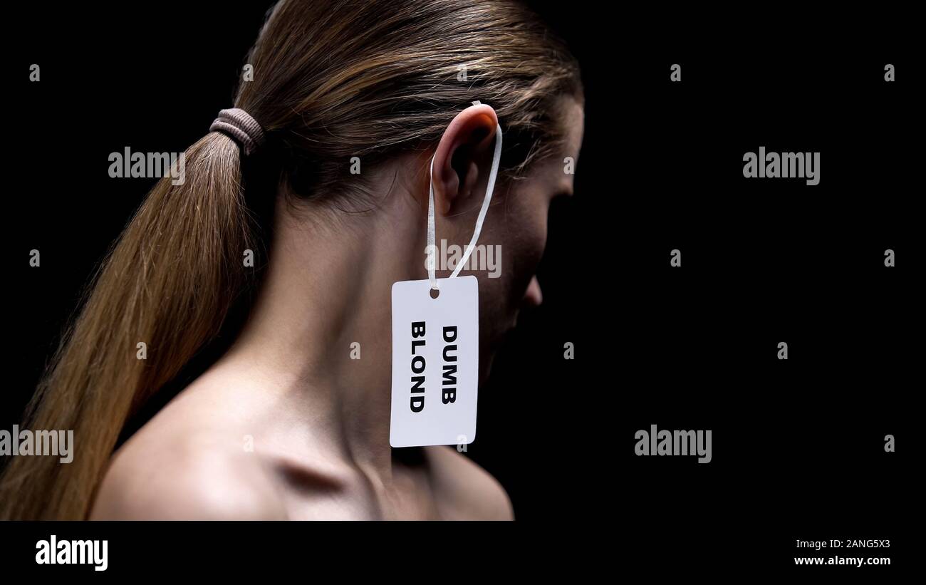 Lady with dumb blonde tag on ear against dark background, humiliation stereotype Stock Photo