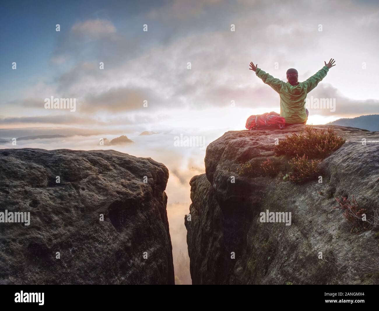 Young woman sit down on top of a mountain celebrate the day. Young lady hiker sitting in blue green windbreaker with red rucksack on sharp cliff Stock Photo