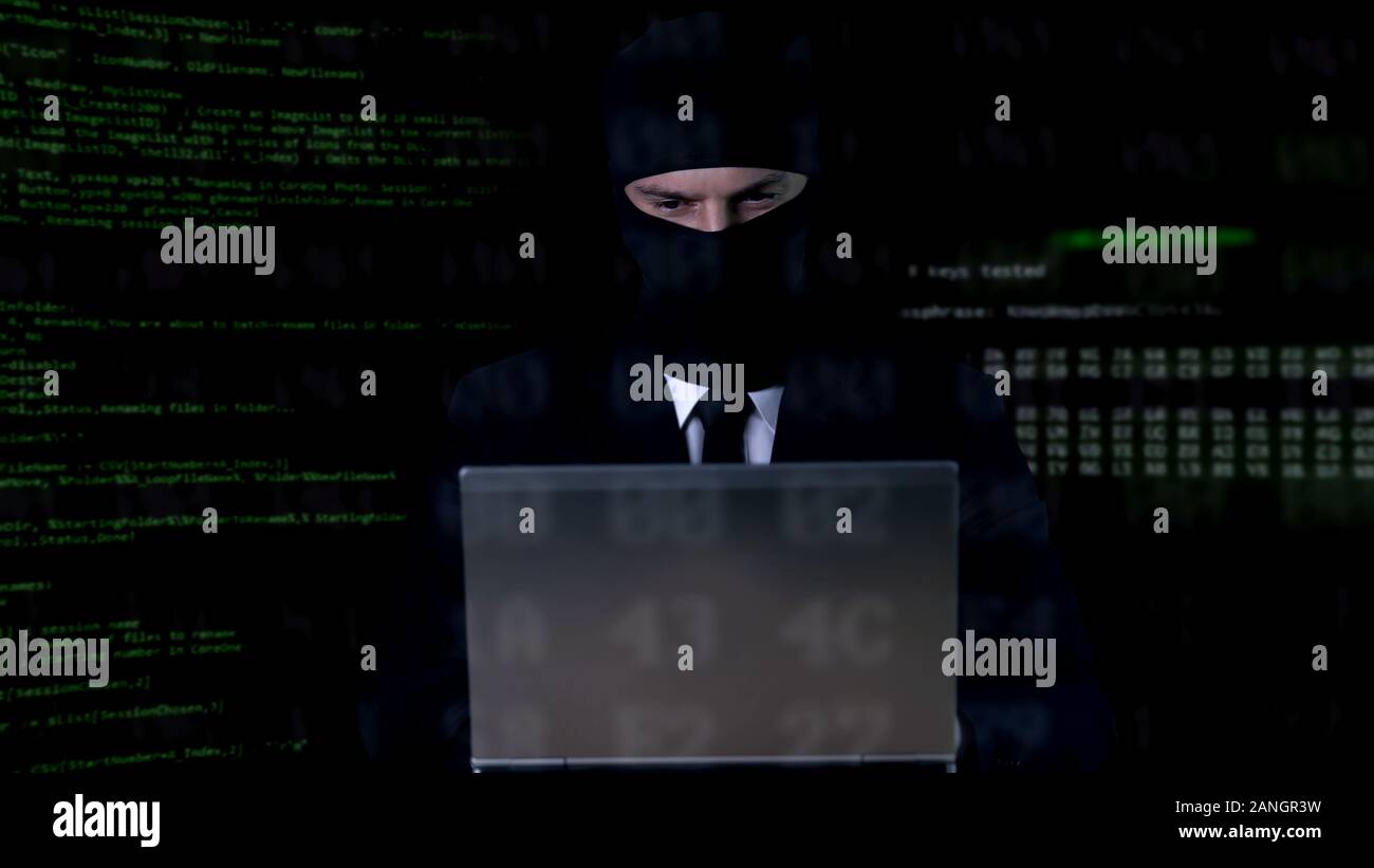 Hacker in balaclava working on laptop, illegal privacy attack, data leakage Stock Photo