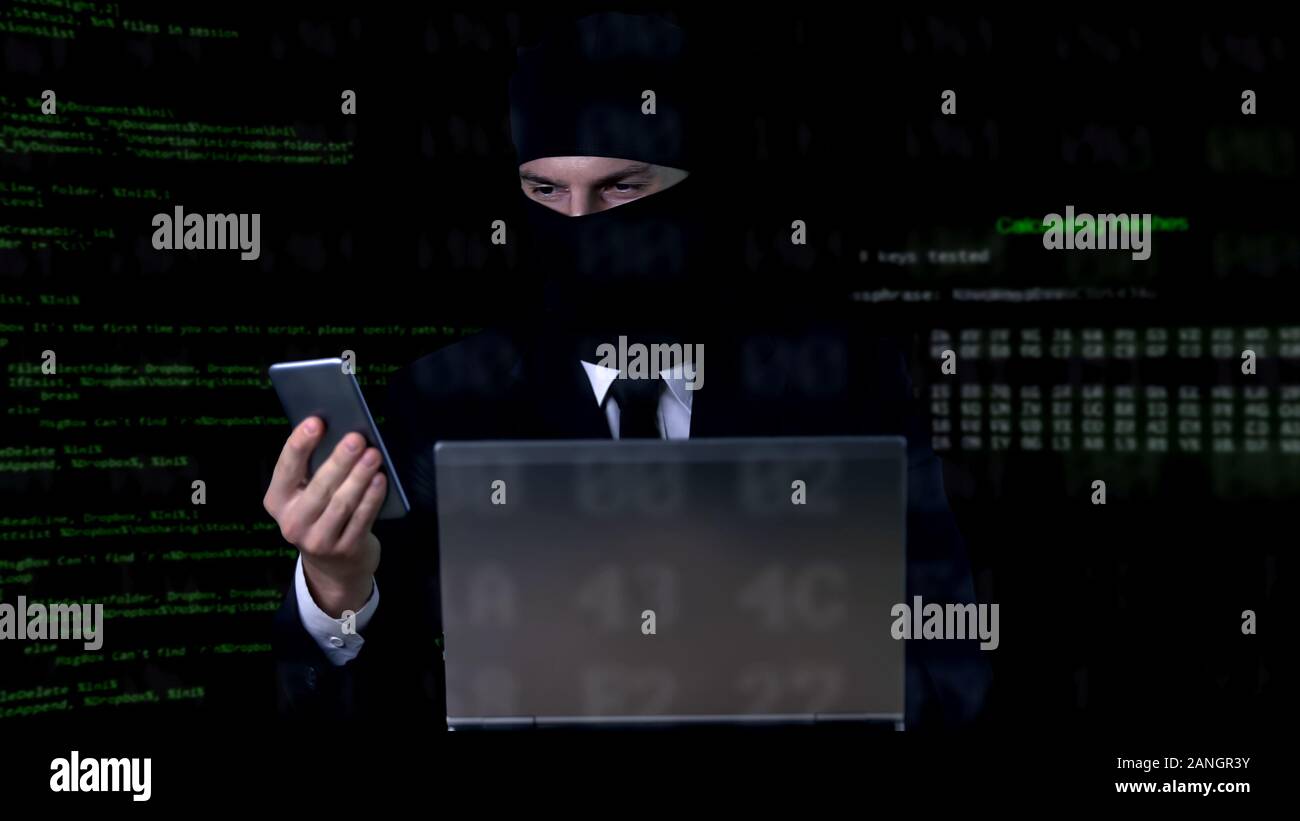 Criminal in suit and balaclava breaking security code using laptop and phone Stock Photo