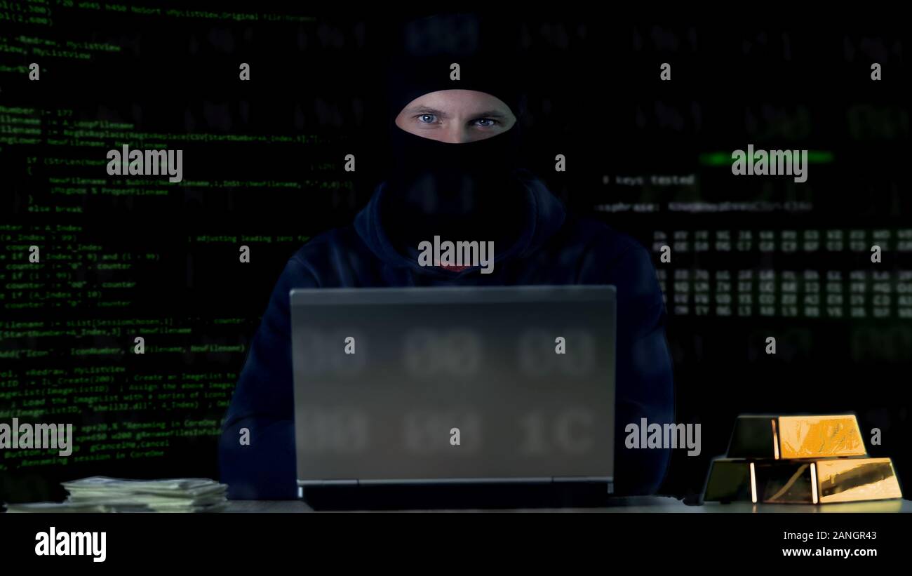 Hacker in balaclava committing cyber crime on laptop, numbers codes background Stock Photo