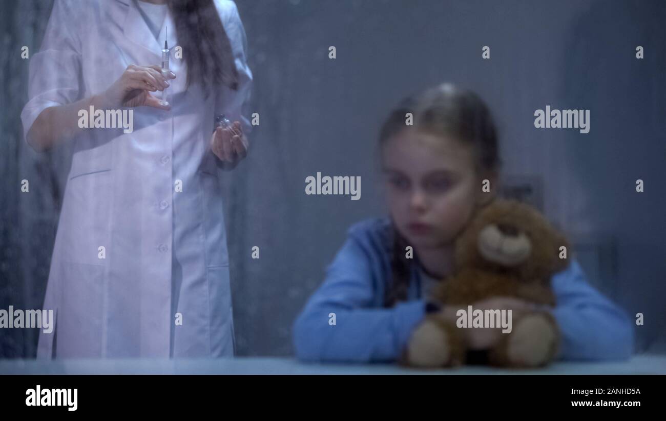 Frightened girl holding teddy bear, nurse with syringe on background, rainy day Stock Photo