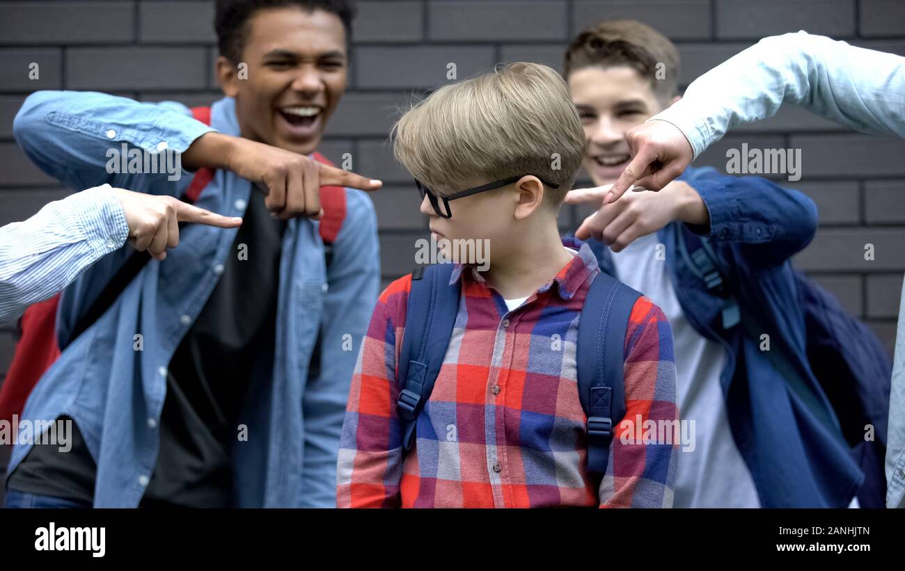 Schoolchildren pointing fingers at junior student, mocking smart boy, bullying Stock Photo