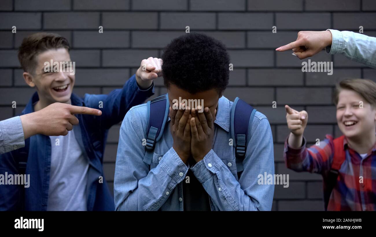 Mean teenagers pointing fingers at black boy crying, verbal bullying, bad rumors Stock Photo